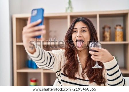 Hispanic young business woman taking a selfie picture drinking a glass of wine sticking tongue out happy with funny expression. 