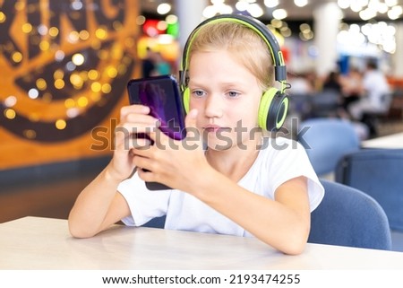A little girl with headphones is watching cartoons on her phone sitting at a table in a cafe. Cute schoolgirl listening to music with mobile phone and headphones. Education, lifestyle
