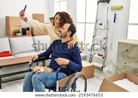 Middle age hispanic couple smiling happy. Man sitting on wheelchair with dog on his legs and woman making selfie by the smartphone at new home.