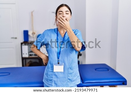 Young hispanic woman wearing physiotherapist uniform standing at clinic bored yawning tired covering mouth with hand. restless and sleepiness. 