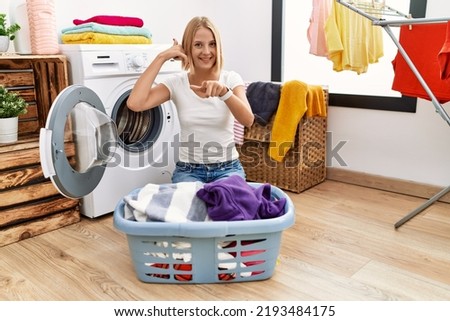 Young caucasian woman doing laundry with clothes in the basket smiling doing talking on the telephone gesture and pointing to you. call me. 