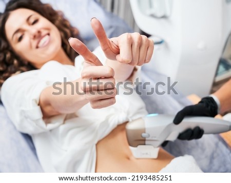 Young satisfied patient with joyful face expression showing thumbs up during SMAS - lifting session in aesthetic medicine clinic. Non-operative method rejuvenation body skin. Focus on female hands. Royalty-Free Stock Photo #2193485251