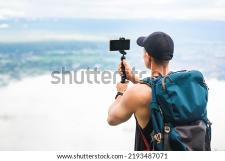Back view of tourist man with backpack using smart phone on selfie stick shooting video blog. Asian traveler taking photo on vacation trip with beautiful sky on background. Royalty-Free Stock Photo #2193487071