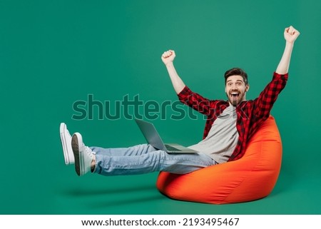 Full body young happy man he 20s wearing red shirt grey t-shirt sit in bag chair hold use work on laptop pc computer do winner gesture isolated on plain dark green background People lifestyle concept