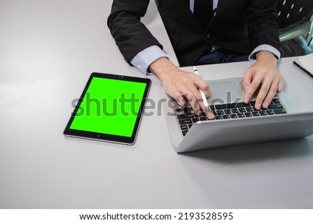 the businessman uses a laptop and holds a digital pen for writing on a green screen tablet, sitting on the table in the modern office for working and learning about technology
