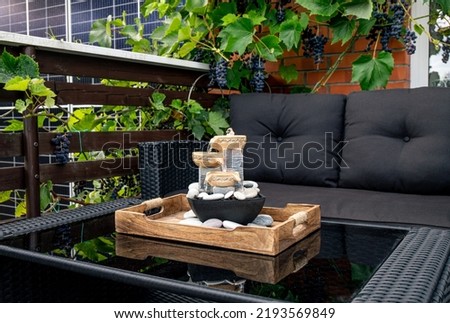 Home patio with black plastic garden furniture, small relaxing electrical zen table fountain on table and real grape vines with grapes hanging on background. Home decor. Royalty-Free Stock Photo #2193569849