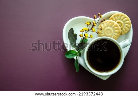 Flatlay A cup of coffee and two small round delicious cookies on a saucer in the shape of a heart lies a twig with small yellow flowers and leaves in the lower corner, Flat lay white on a dark purple 