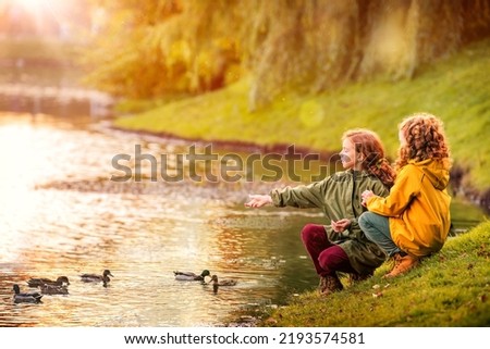 Two red-haired schoolgirl girls, sisters, cheerfully feeding ducks on bank of a pond in a city park during the golden autumn leaf fall.