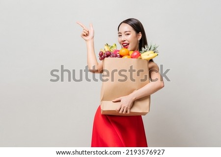 Pretty Asian woman in red dress holding paper grocery bag full of tropical fruits pointing hand up in gray studio isolated background