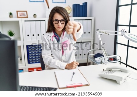 Young doctor woman wearing doctor uniform and stethoscope at the clinic looking unhappy and angry showing rejection and negative with thumbs down gesture. bad expression. 