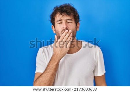 Hispanic young man standing over blue background bored yawning tired covering mouth with hand. restless and sleepiness. 