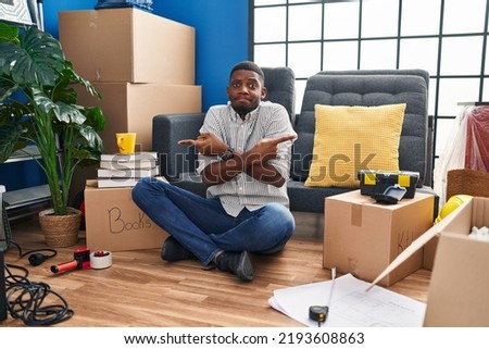 African american man sitting on the floor at new home pointing to both sides with fingers, different direction disagree 