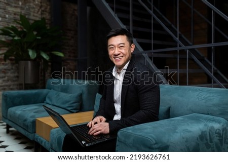 Portrait of successful Asian businessman, man working on laptop smiling and looking at camera, boss in business suit sitting on sofa in modern office, investor happy satisfied