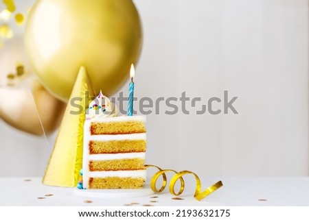 Slice of birthday cake with single blue birthday candle and gold birthday balloons