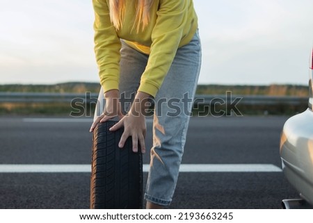 girl in stands near the rear wheel of the car and rolls a spare wheel to change it.A car breakdown, the wheel in front is a temporary stop sign blurred on the right there is a place for an