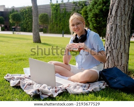 A young student girl in glasses sits in a park on the grass, uses a laptop, eats a sandwich.