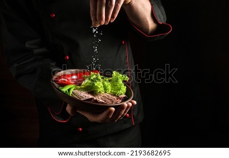 Professional chef sprinkles salt on a sliced steak with beef and vegetables in a plate. The concept of serving dishes to order