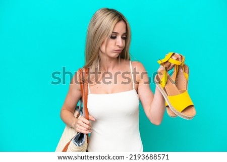 Blonde woman in swimsuit holding summer sandals isolated on blue background with sad expression