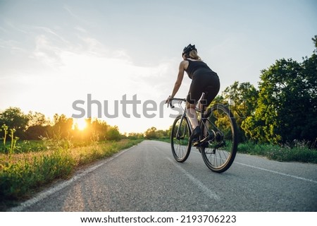Rear view of a young woman cyclist riding road bicycle on a free road in the nature at a sunny day. Healthy lifestyle concept. Riding into the sunset. Copy space. Royalty-Free Stock Photo #2193706223