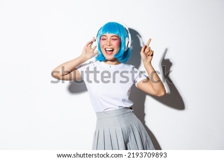 Happy beautiful japanese girl in blue wig listening music in headphones, dancing and singing along, standing over white background