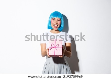 Image of cheerful cute asian girl in blue party wig, giving you a gift wrapped in pink paper, congratulating with something, standing over white background