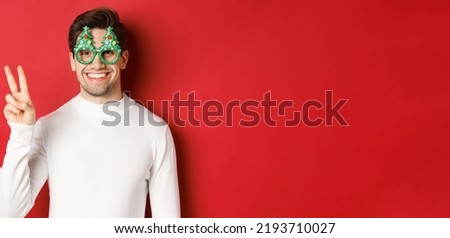 Image of handsome man in white sweater and party glasses, showing peace sign and smiling, wishing merry christmas, standing over red background