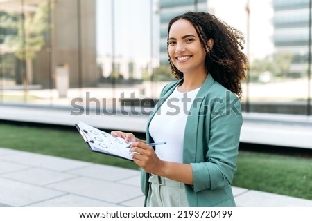 Portrait of a successful smart business woman, brazilian or hispanic nationality, financial consultant, mentor, stands outdoors, holding financial documents in her hand, looking at the camera