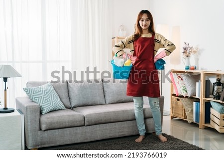 full length professional asian female housekeeper is standing and carrying a bucket of cleaning tools while smiling at the camera in a tidy living room interior