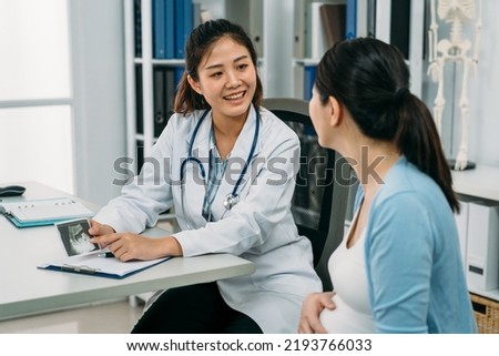 asian female gynecologist is smiling while showing and explaining an ultrasound picture to her expectant patient at the office desk in the clinic.