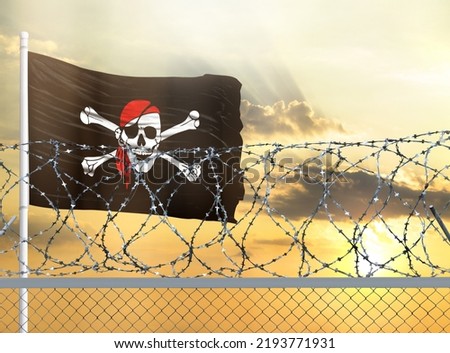 Flagpole with the flag of Jolly Roger pirate against the sky and behind a fence with barbed wire. The concept of protecting the borders of territories.