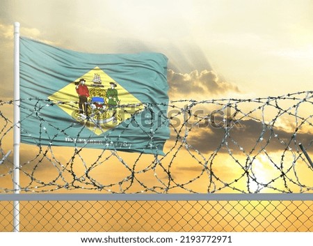 Flagpole with the flag of State of Delaware against the sky and behind a fence with barbed wire. The concept of protecting the borders of territories.