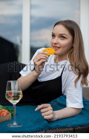 Portrait of young happy attractive woman wearing white T-shirt, black sarafan, lying on blue blanket near glass of white wine, cut fruits on wooden tray, eating slice of pepper on window background.