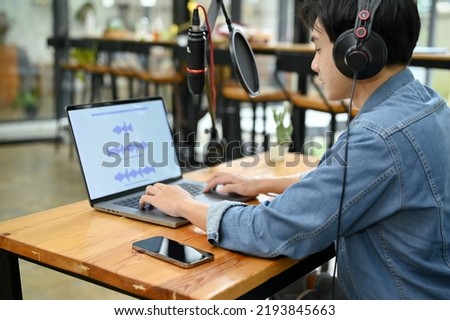 Talented young Asian male radio host running his online radio channel in her studio, adjusting the radio sounds in his laptop computer.