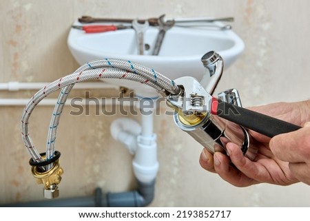 Plumbing work on installing water faucet on sink in bathroom, plumber fixes braided connecting hoses on tap using adjustable wrench. Royalty-Free Stock Photo #2193852717