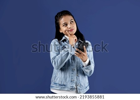 Potrait of young asia woman holds mobile phone has thoughtful expression thinks about received message wears denim jacket looks away isolated over blue background.