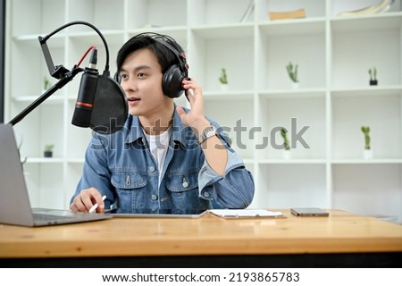 Talented and smart young Asian male radio host or VJ wearing headphones and speaking on his professional microphone, running his podcast in studio.
