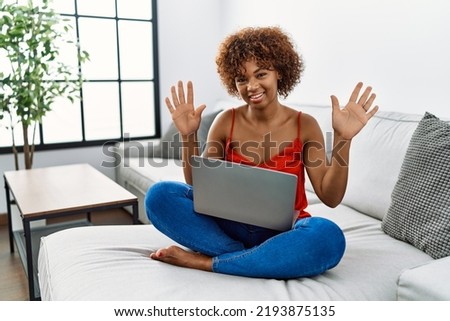 Young african american woman sitting on the sofa at home using laptop showing and pointing up with fingers number ten while smiling confident and happy. 