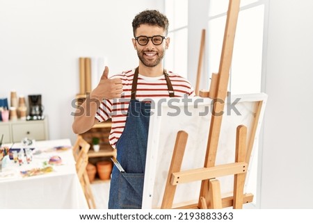 Arab young man at art studio doing happy thumbs up gesture with hand. approving expression looking at the camera showing success. 