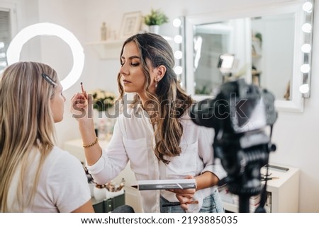 Happy young woman streaming a beauty makeup vlog from home or workshop. Beautiful online content creator cosmetician applying makeup and explaining some work tools. Vlogging and online channel work.  Royalty-Free Stock Photo #2193885035