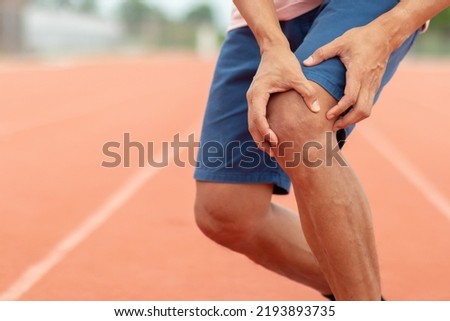Picture of knee pain of a 45-year-old man with both hands cradling his left knee. In the red running track at the stadium Because the muscles are inflamed from running too hard.