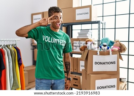 Young handsome hispanic man wearing volunteer t shirt at donations stand doing peace symbol with fingers over face, smiling cheerful showing victory 