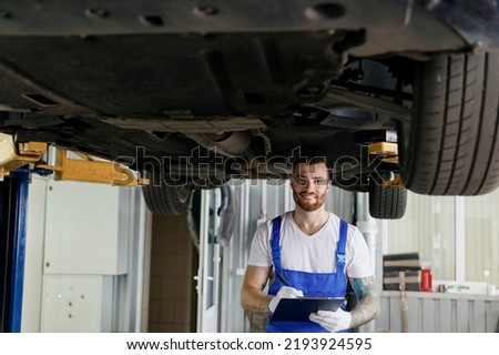 Technician man in denim overalls use hold clipboard papers document writing estimate outlay stand near car lift check technical condition work in vehicle repair workshop indoor. Tattoo translate fun