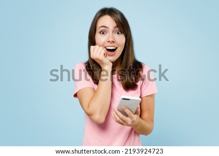 Young surprised happy caucasian woman 20s in pink t-shirt look camera hold in hand use mobile cell phone prop up chin isolated on pastel plain light blue background studio. People lifestyle concept. Royalty-Free Stock Photo #2193924723