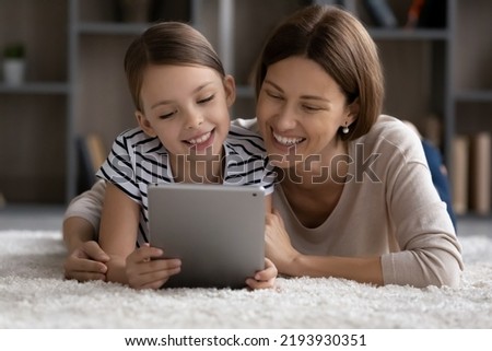 Smiling young mother cuddling adorable kid daughter, using digital tablet together at home lying on fluffy floor carpet, enjoying watching funny cartoons or photo video content, playing games online