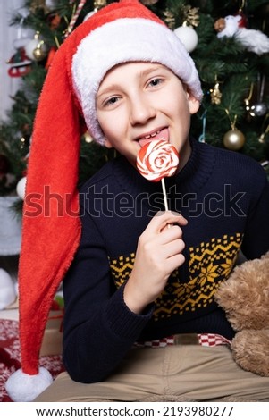 A boy in a Santa Claus hat licks a lollipop on a stick near a decorated Christmas tree.