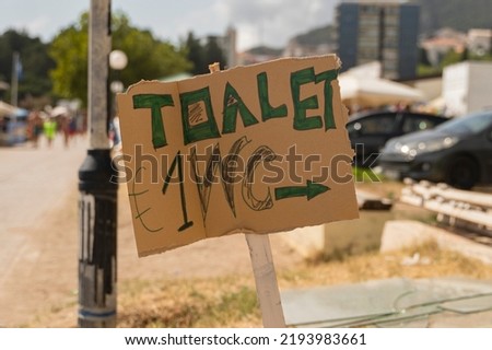 Close-up of a cardboard sign that shows the direction towards a public private toilet. The cost of a public toilet in the resort town is 1 euro sign.