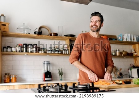 Stock photo of a middle aged man holding a knife and slicing up some vegetables in a kitchen. He is looking through a window and smiling. Royalty-Free Stock Photo #2193989175