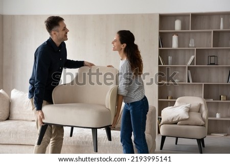 Happy beautiful hispanic young family couple homeowners holding armchair, enjoying renovating living room rearranging furniture together. Smiling spouses relocating on moving day into own dwelling. Royalty-Free Stock Photo #2193995141