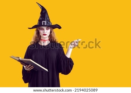 Serious creepy woman in witch character pointing with finger to side on orange background in studio. Portrait of woman with bloody makeup in black dress and witch's hat holding spell book. Web Royalty-Free Stock Photo #2194025089