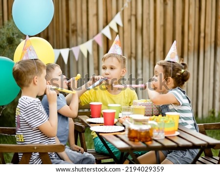 Cute funny nine year old boy celebrating his birthday with family or friends with homemade baked cake in a backyard. Birthday party. Kids wearing party hats and blowing whistles